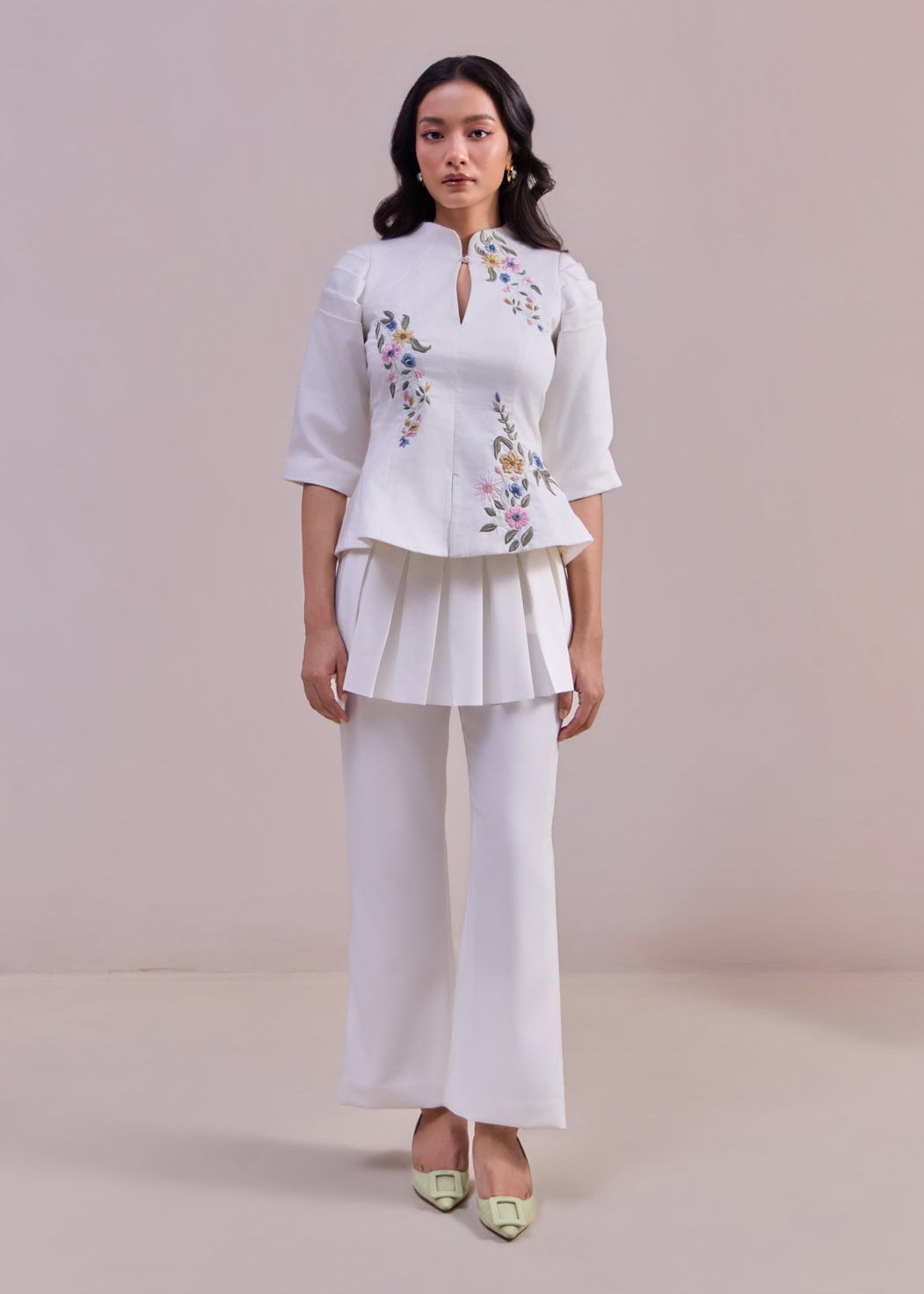 Woman wearing a white blouse with floral embroidery and white pants against a plain background