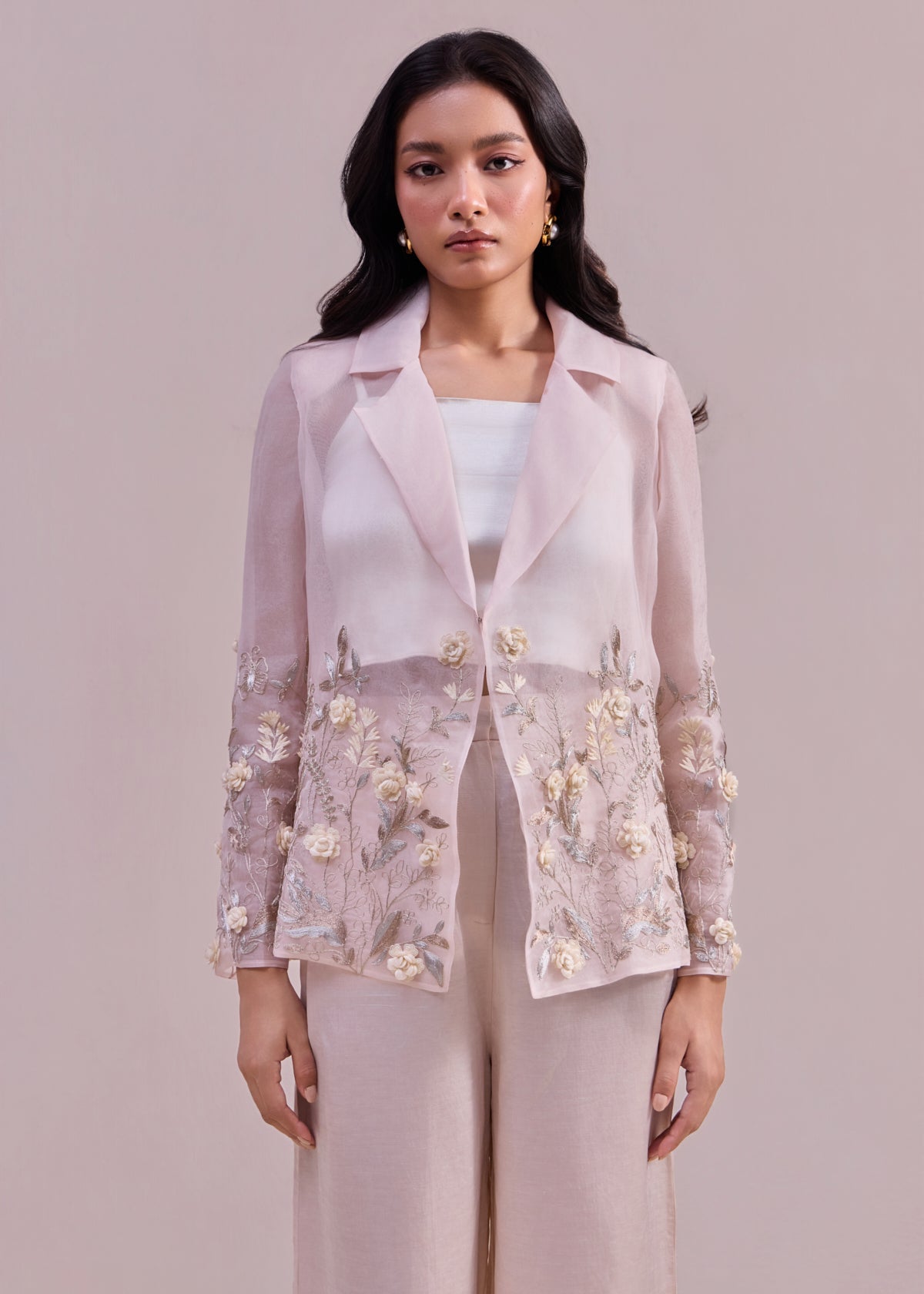 Woman wearing a light pink embroidered blazer over a white top and pants on a plain background