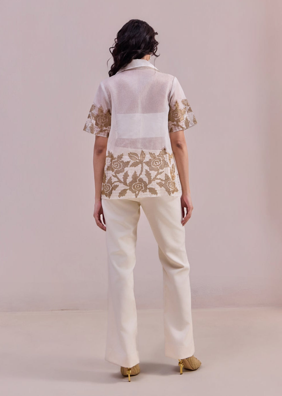 Woman wearing a white blouse with floral embroidery and cream pants on a plain background