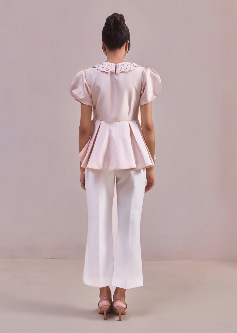 Person wearing a light pink blouse with ruffled sleeves and white pants on a plain background