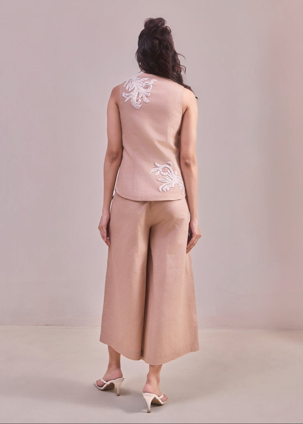 Woman wearing a beige sleeveless top and wide-leg pants with floral embroidery on a plain background