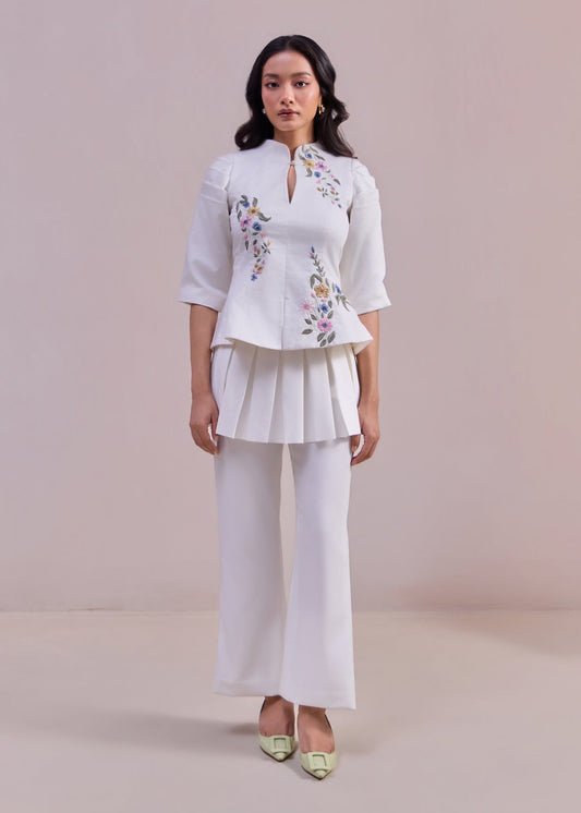 Woman wearing a white blouse with floral embroidery and white pants against a plain background
