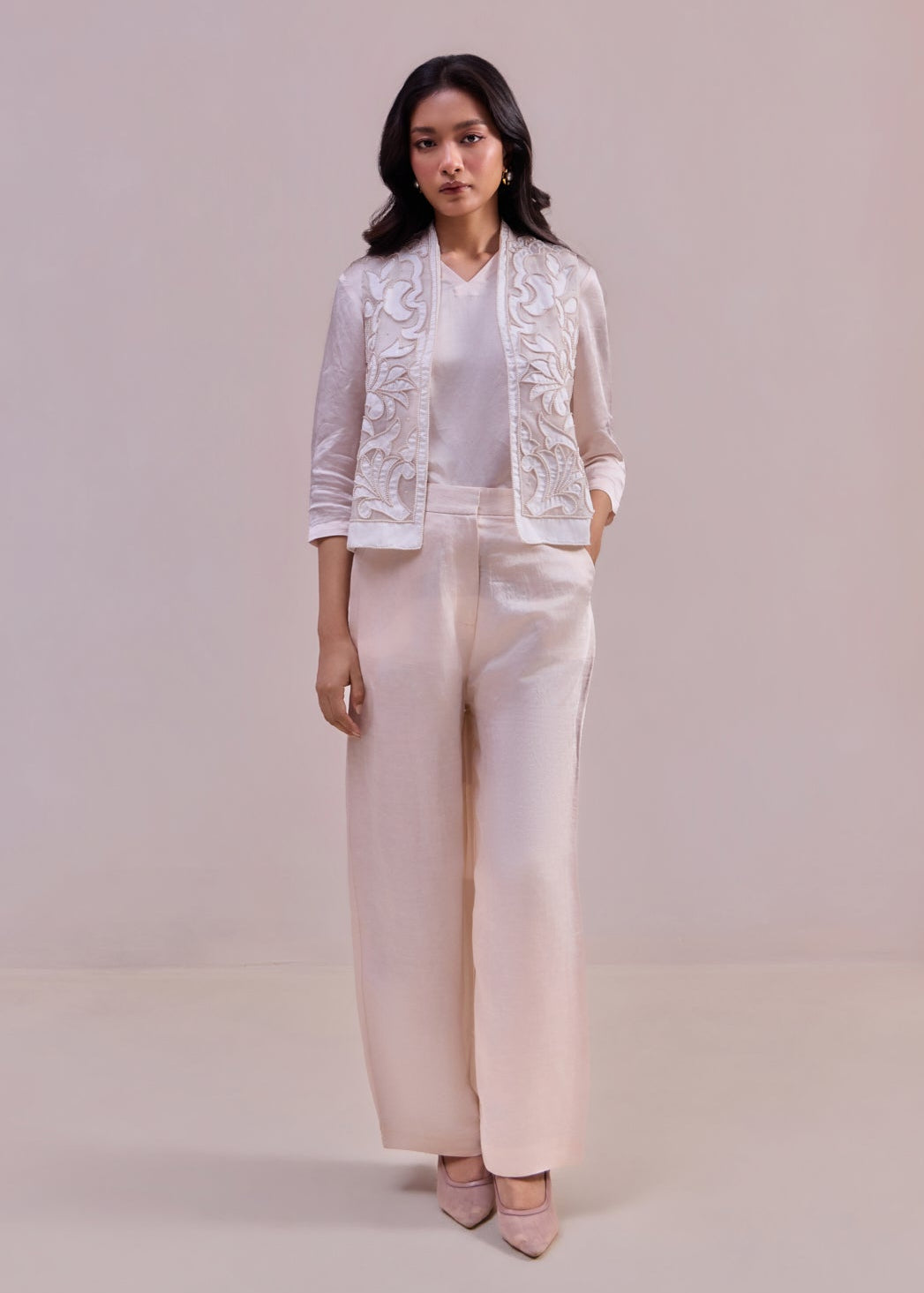 Woman wearing a white lace jacket and light pink pants on a plain background