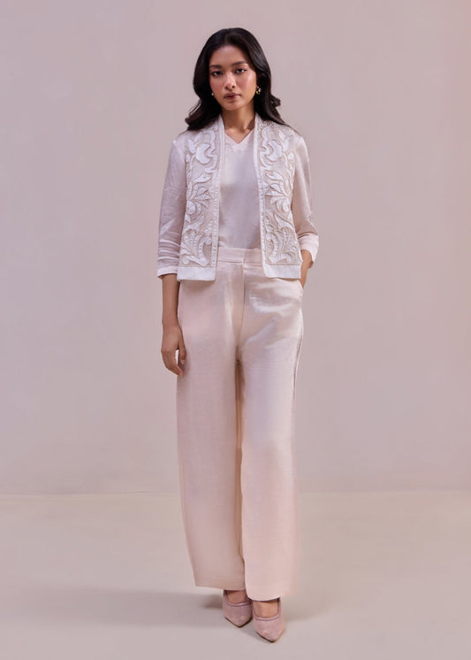 Woman wearing a white lace jacket and light pink pants on a plain background