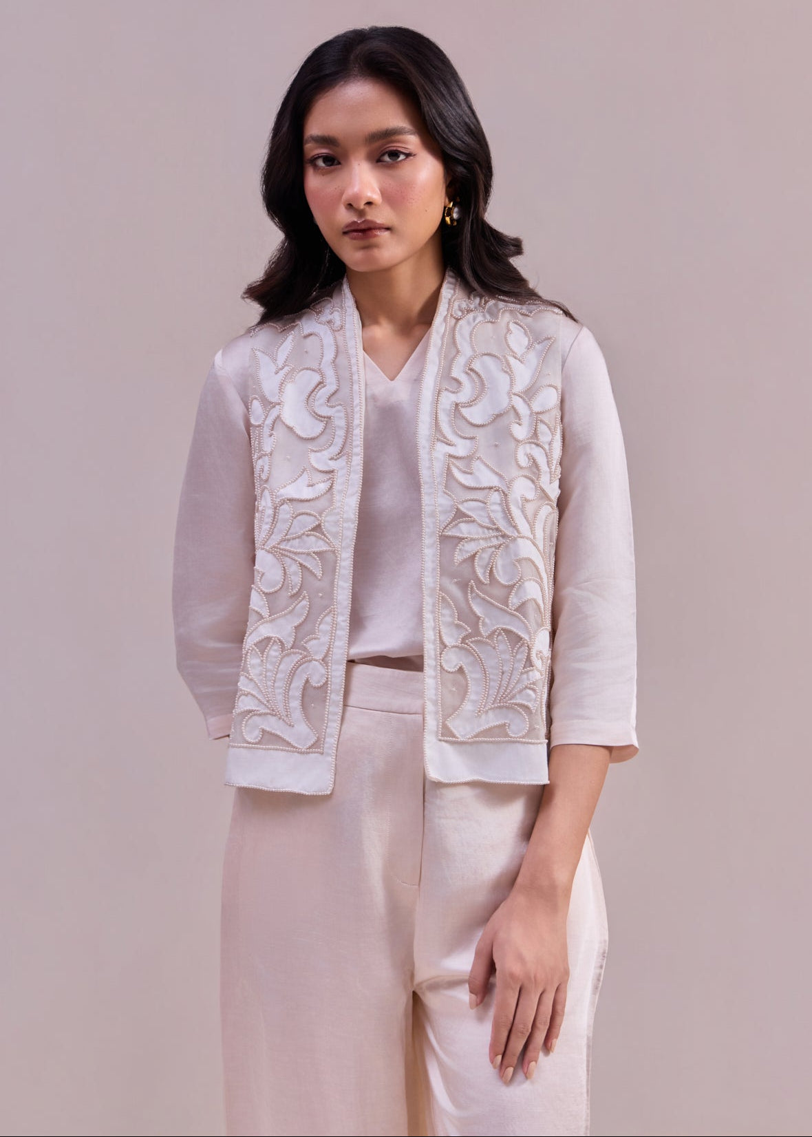 Woman wearing a white embroidered jacket over a white shirt and pants on a beige background