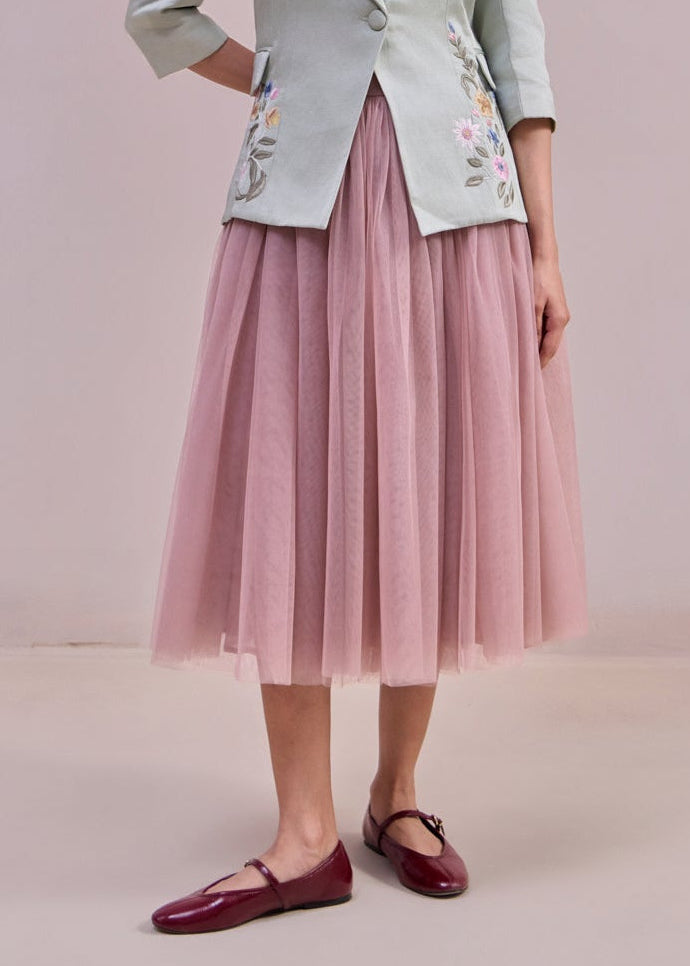 Woman wearing a light green blazer with floral embroidery and a pink skirt against a plain background