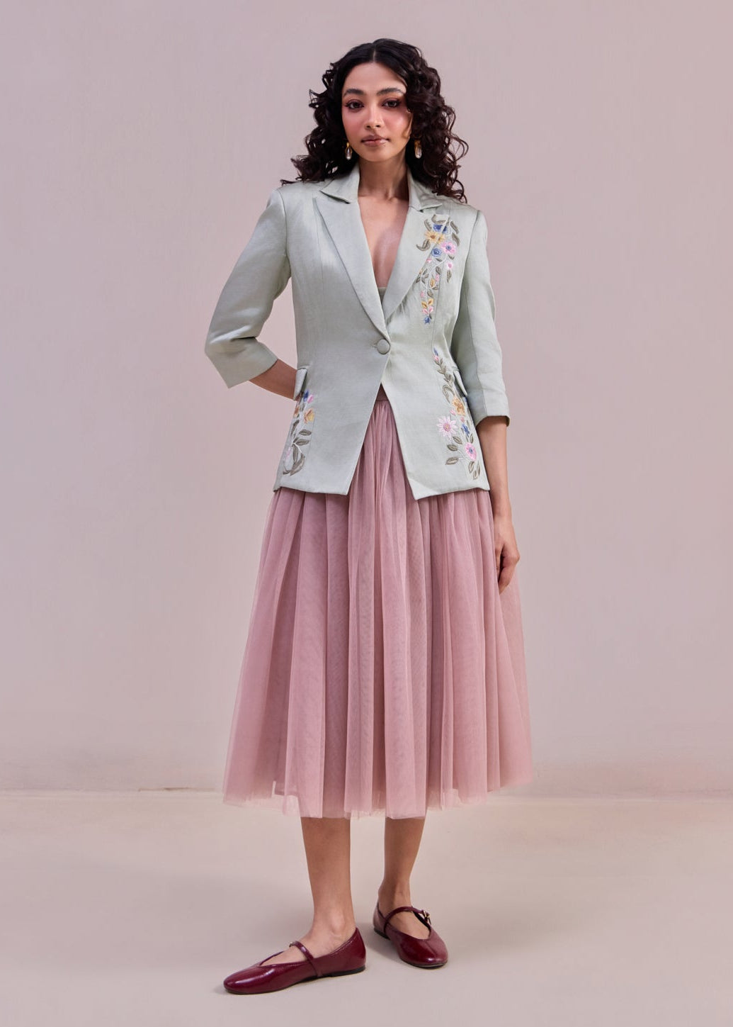 Woman wearing a light green blazer with floral embroidery and a pink skirt against a plain background