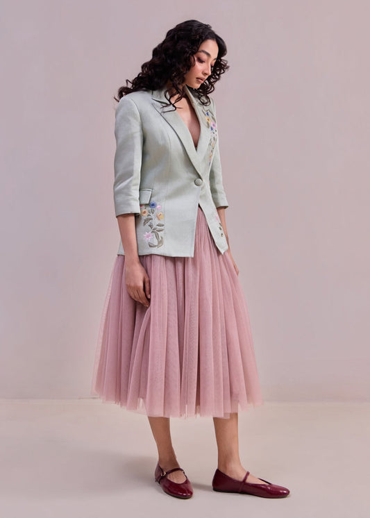 Woman wearing a light green blazer with floral embroidery and a pink tulle skirt on a plain background