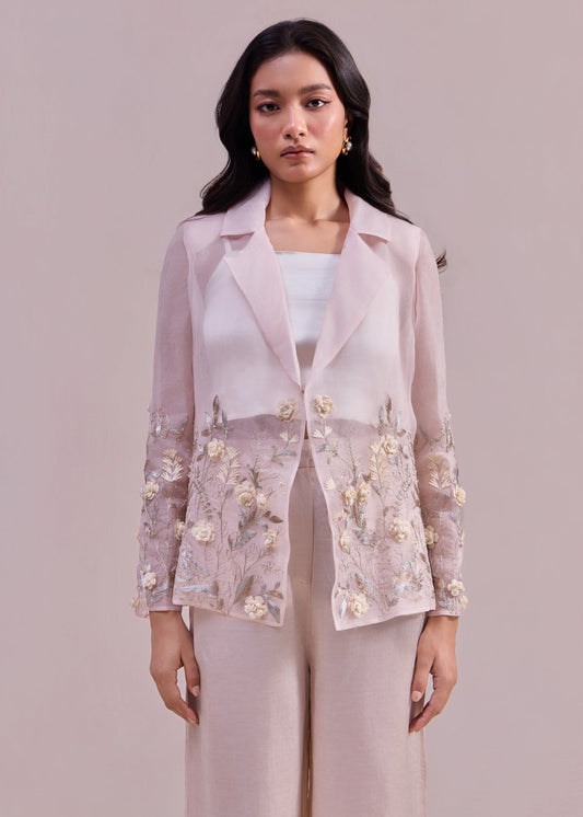 Woman wearing a light pink embroidered blazer over a white top and pants on a plain background