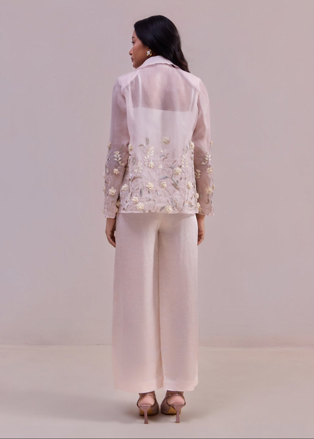Woman wearing a light pink embroidered jacket and pants on a plain background