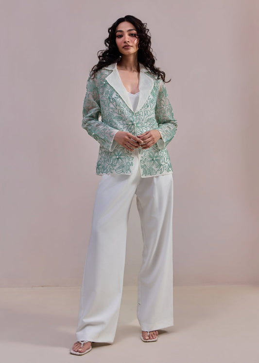 Woman wearing a green patterned blazer and white pants against a plain background