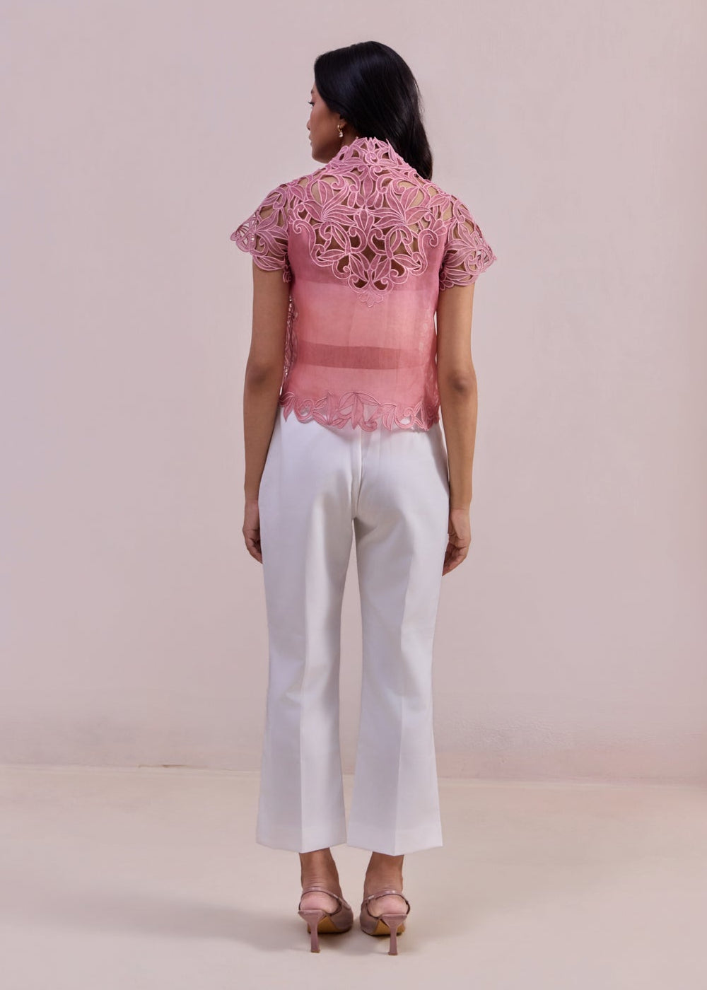 Woman wearing a pink embroidered top and white pants against a plain background