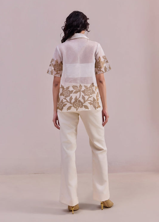 Woman wearing a white blouse with floral embroidery and cream pants on a plain background
