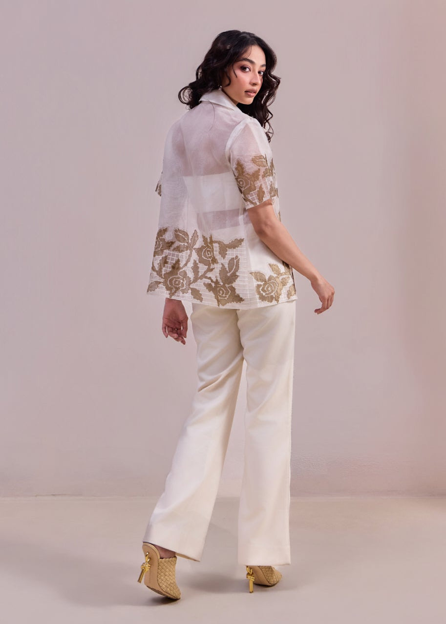 Woman wearing a white floral blouse and cream pants against a plain background