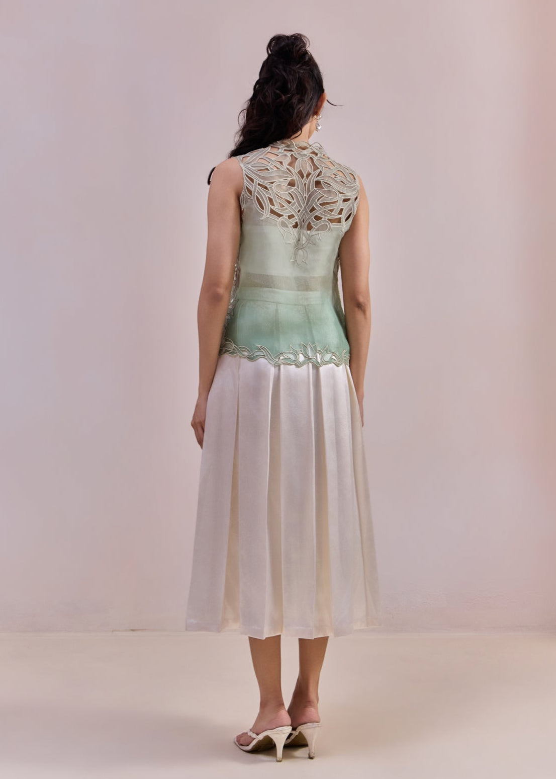 Woman wearing a light green sleeveless top with lace details and white pants on a plain background