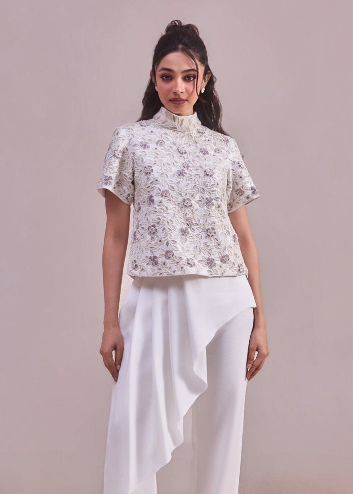 Woman wearing a floral blouse and white pants against a plain background