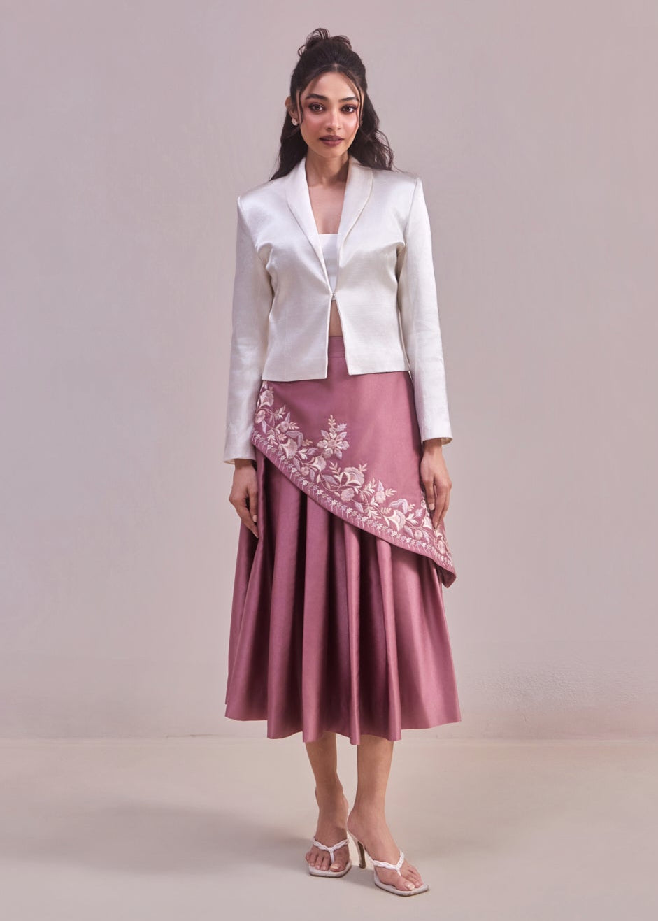 Woman wearing a white blazer and pink skirt with floral embroidery on a plain background