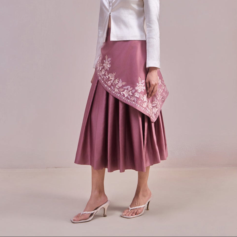 Woman wearing a white blouse and pink skirt with floral embroidery against a plain background