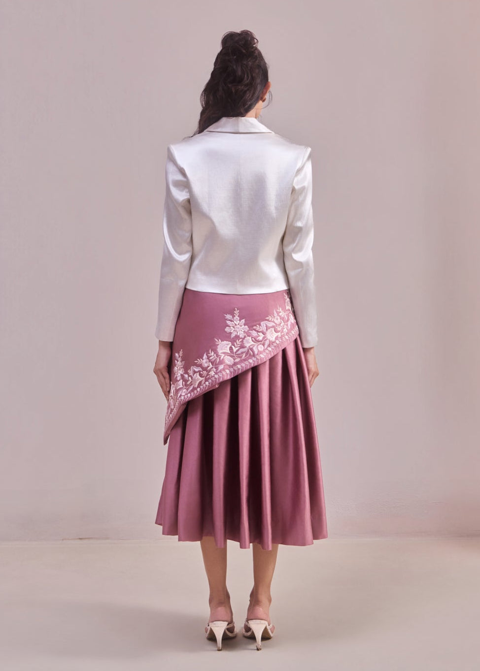 Woman wearing a white jacket and pink skirt with floral embroidery on a plain background