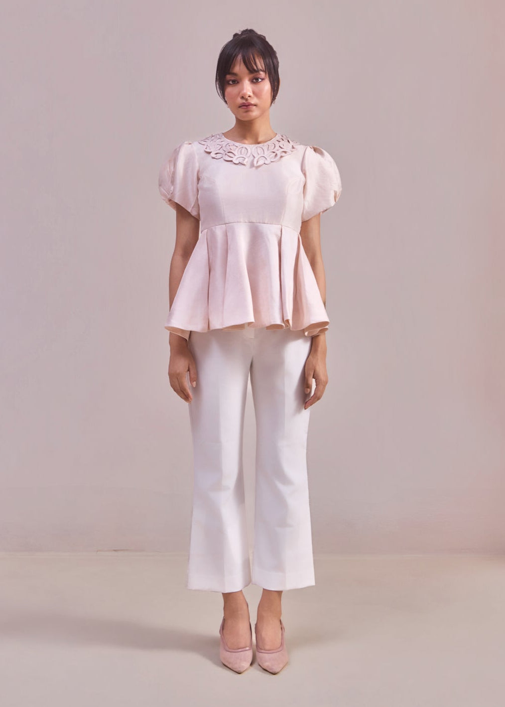 Woman wearing a light pink blouse with lace details and white pants on a plain background