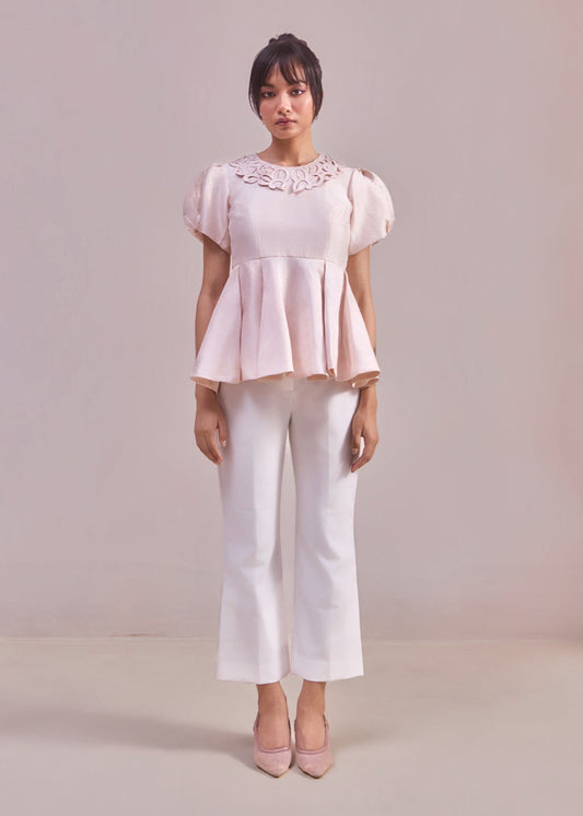 Woman wearing a light pink blouse with lace details and white pants on a plain background