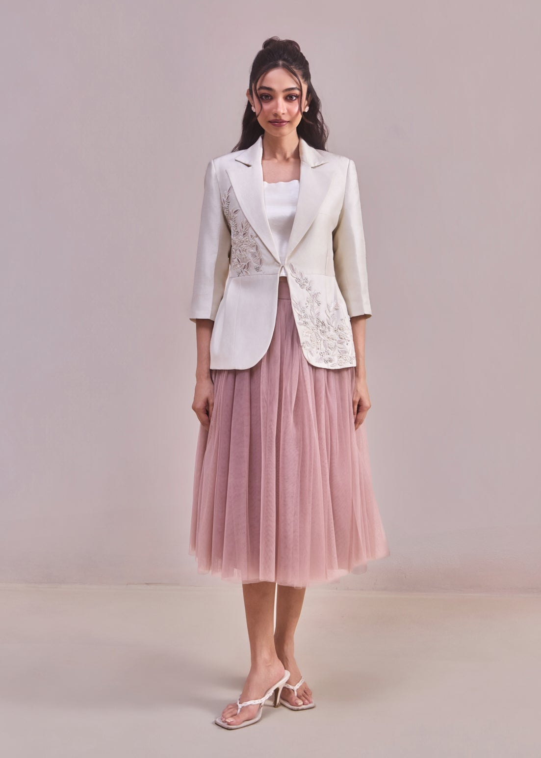 Woman wearing a white blazer with floral embroidery and a pink tulle skirt on a plain background