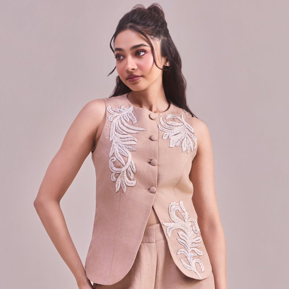 Woman wearing a beige embroidered vest and pants on a light pink background