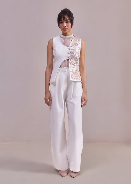 Person wearing a white sleeveless top with floral details and white pants against a plain background