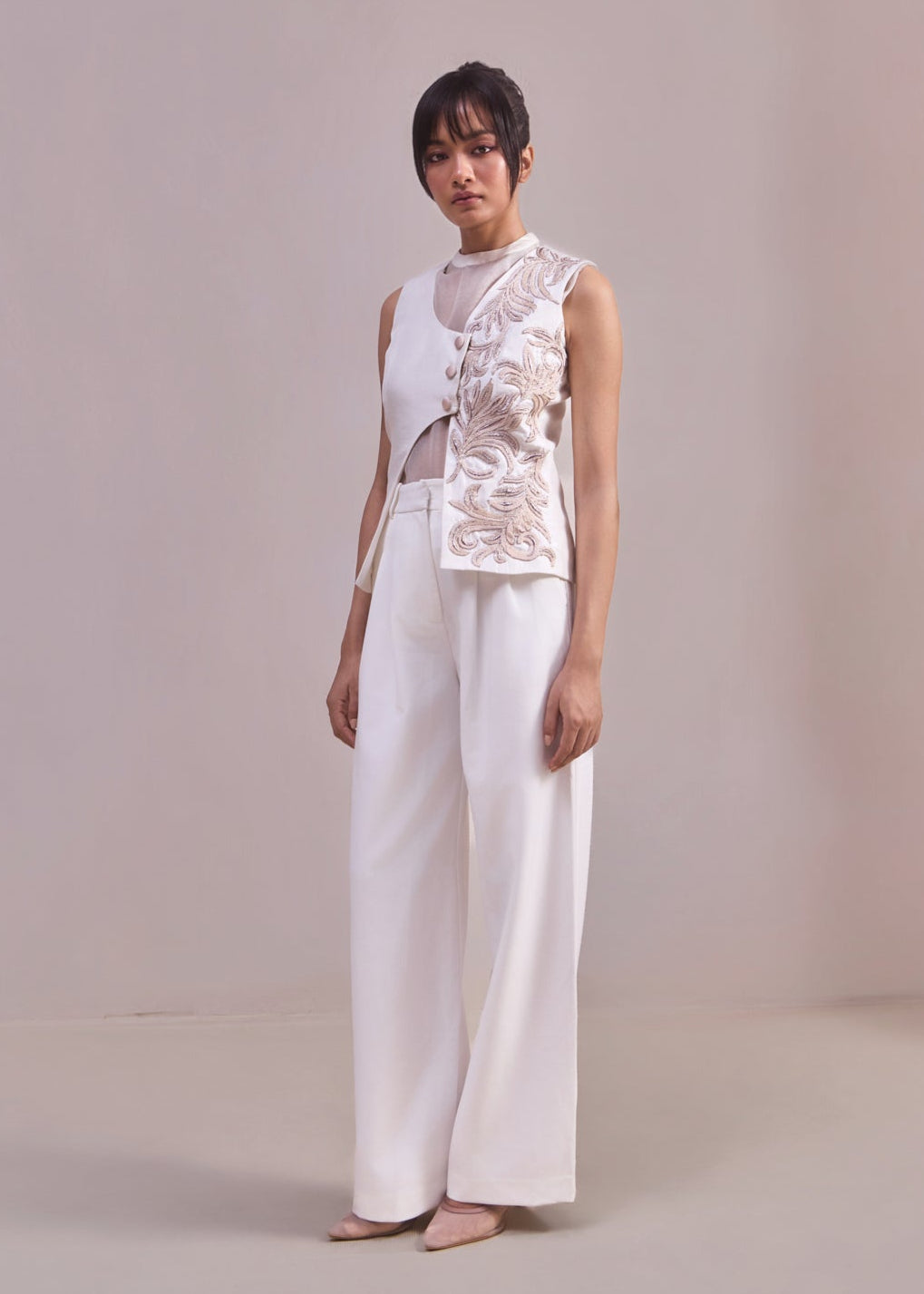 Woman wearing a white sleeveless top with lace details and white pants against a plain background