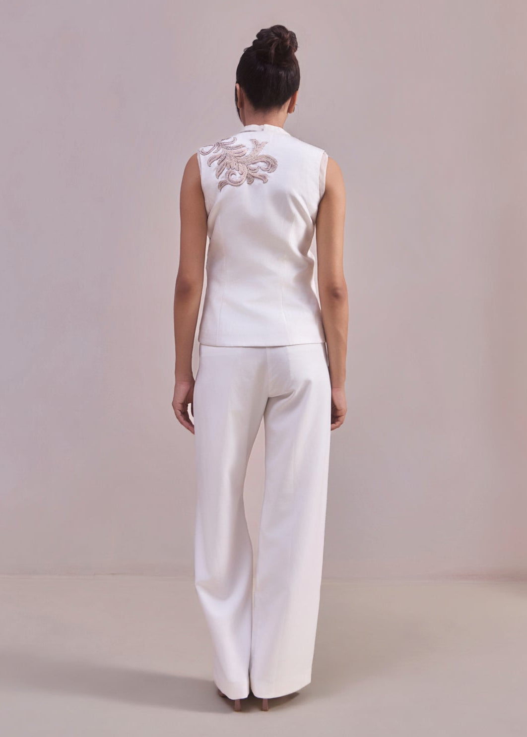 Woman wearing a white sleeveless top and pants with floral embroidery on a plain background